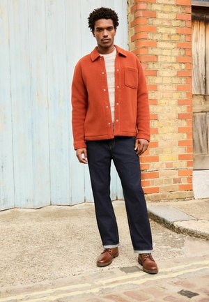 Orangefarbene Fleece-Hemdenjacke mit Druckknöpfen, darunter ein grau gestreifter Pullover, dunkelblaue Jeans und braune Schnürschuhe. Texturierte Stoffdetails.