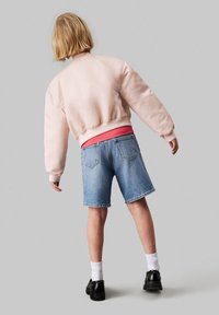 Chaqueta bomber rosa claro con ribetes en el dobladillo, usada sobre una camiseta roja. Shorts de mezclilla azul con un corte holgado y dobladillos enrollados, zapatos negros, calcetines blancos.