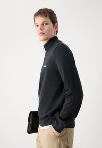 Sudadera de forro polar negra con cierre, de acabado texturizado, con un cuello alto y un logo sutil. Combinada con pantalones caqui y un bolso negro.