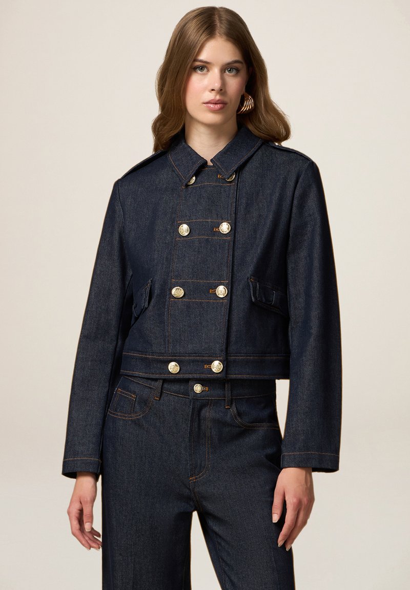 Jachetă din denim, croită, de culoare albastru închis, cu guler structurat, butoni argintii cu două rânduri și detalii de cusătură portocalie.