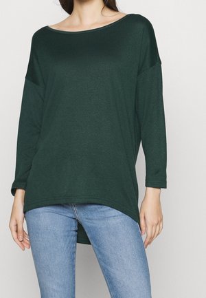 Kobieta ubrana w ciemnozielony, oversize'owy top z długim rękawem i dekoltem typu łódka oraz jasnoniebieskie jeansy na jednolitym tle.