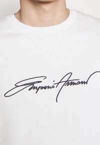 Vit sweatshirt med rund hals, som har en framträdande svart broderad signatur över bröstet. Mjukt tyg med en slät textur.