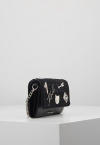 Piccola borsa nera con pattina in tweed adornata da spille in metallo tra cui un gatto, una macchina fotografica, una freccia e una tracolla a catena, posizionata su una superficie bianca.