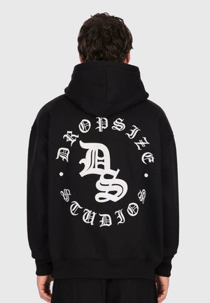 Persona che indossa un hoodie nero con grande testo bianco in stile gotico "Dropsine Studios" e il logo stilizzato "DS" sulla schiena.