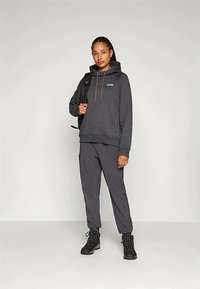 Žena stoji i nosi tamnu prešanu kapu, usklađene trenirke, crne planinarske čizme i nosi crni ruksak prebačen preko jednog ramena.