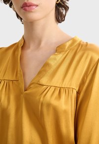 Cuello y hombro de una persona con cabello corto castaño, que lleva un pendiente de aro dorado y una blusa brillante de color mostaza con escote en V.