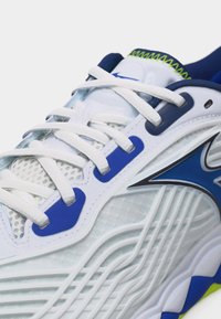 Λευκό και μπλε αθλητικό παπούτσι με λευκά κορδόνια, ανώτερο μέρος από διχτυωτό ύφασμα, υφή στη σόλα και ραφές νέον πράσινου χρώματος στη γλώσσα και στον γιακά.