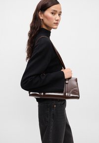 Brun skinn skulderveske med glanset overflate, bånddesign og en lang stropp, holdt av en person i svart genser og jeans.