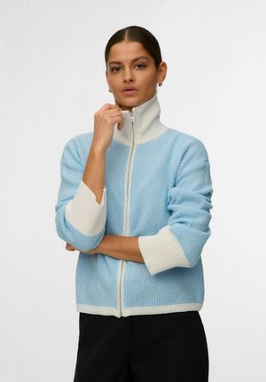 Kvinne med mørkt hår iført en lyseblå og hvit genser med glidelås, holder glidelåsen nær halsen, stående mot en ensfarget bakgrunn.