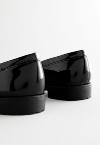 Chaussures en cuir verni noir avec un profil bas, un embout arrondi et une semelle en caoutchouc texturée ; présentent une finition brillante et un détail de couture contrastante.