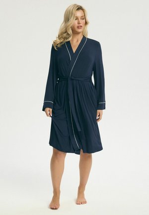 Kobieta o blond włosach, ubrana w granatowy szlafrok do kolan z białym paskiem i przewiązanym pasem, stojąca boso na jednolitym białym tle.