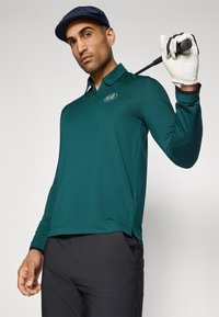 Muškarac u tamnozelenoj dugoj majici polo i crnim hlačama drži golf štap preko ramena s bijelom rukavicom i mornarskom kapom protiv bijele pozadine.