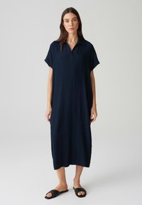 Abito blu navy con collo rialzato, maniche corte e vestibilità ampia. Il tessuto testurizzato arriva a metà polpaccio, abbinato a sandali neri a scivolo.