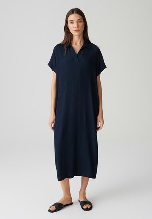 Abito blu navy con collo rialzato, maniche corte e vestibilità ampia. Il tessuto testurizzato arriva a metà polpaccio, abbinato a sandali neri a scivolo.