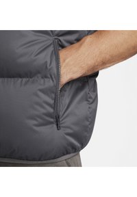 Gilet grigio imbottito con tasca laterale con zip. Il tessuto appare liscio, con una finitura opaca sottile e cuciture a contrasto lungo le cuciture.