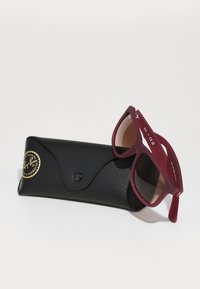 Ochelari de soare Ray-Ban cu un cadru din cauciuc vișiniu și lentile maronii, așezați pe un etui texturat negru cu un logo auriu.