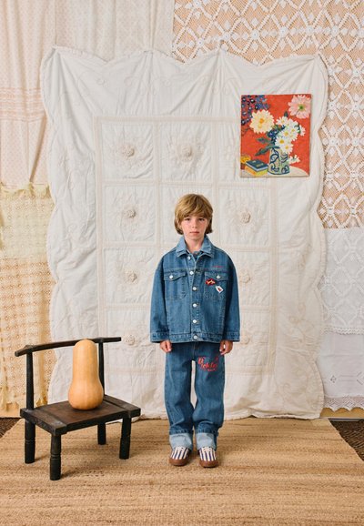Enfant aux cheveux blonds debout, portant une veste en denim et un jean, devant un tissu matelassé blanc avec une peinture florale, à côté d'une chaise en bois tenant une gourde.