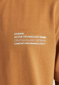 Brun kortærmet skjorte med hvid tekst, der står "CHASIN' ACTIVE-TECHNOLOGY-DENIM SKABER BALANCE MELLEM KOMFORT-YDEEVNE-NYTTE."