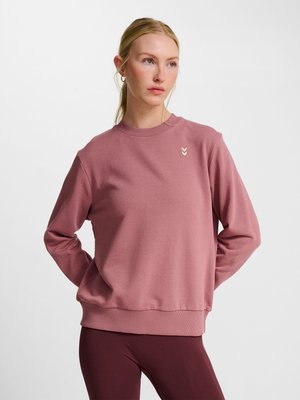 Frau trägt ein staubig rosa langärmeliges Sweatshirt und dunkel mauve Leggings, steht mit den Händen hinter dem Rücken und schaut zur Seite.