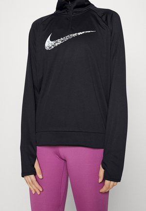 Personne portant un pull Nike noir à manches longues avec un logo blanc et des leggings violets, mains détendues le long du corps sur un fond uni.