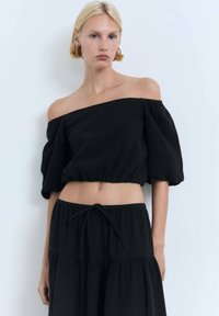 Schwarz, schulterfreies Cropped-Top mit lockeren, gepufften Ärmeln, gerafftem Saum und einem strukturierten Stoff. Kombiniert mit einem passenden, luftigen Rock.
