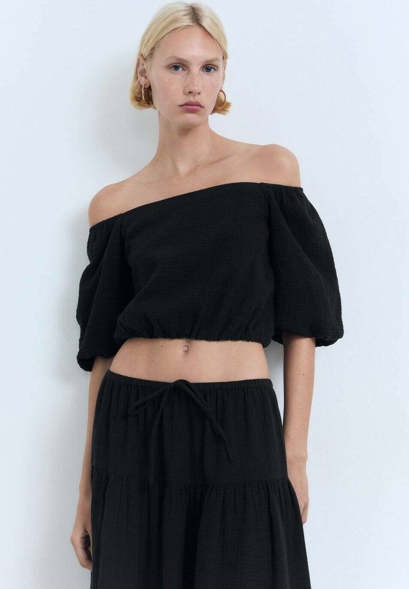 Schwarz, schulterfreies Cropped-Top mit lockeren, gepufften Ärmeln, gerafftem Saum und einem strukturierten Stoff. Kombiniert mit einem passenden, luftigen Rock.