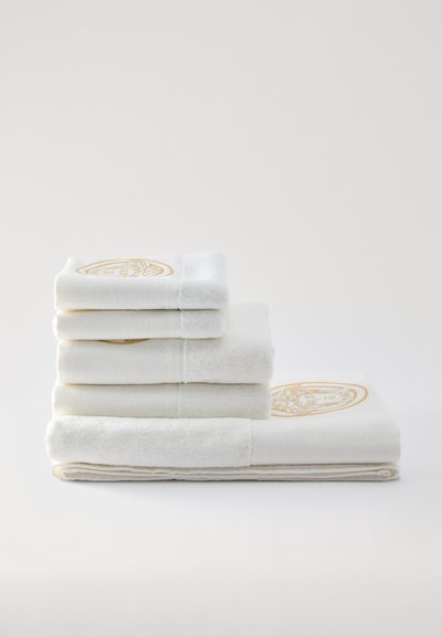 Set de patru prosoape albe stivuite, cu textură netedă de bumbac grosier și emblemă decorativă aurie brodată pe marginea fiecărui prosop.