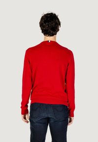Maglione rosso a maglia con maniche lunghe e scollatura rotonda. Presenta polsini e orlo a costine. Dettaglio distintivo sulla parte posteriore del collo con una piccola bandiera.