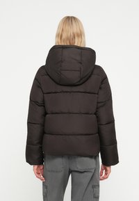 Černá péřová bunda s kapucí, která má prošívaný design, široké rukávy a měkkou, matnou texturu. Pohled zezadu ukazuje odolné detaily švů.