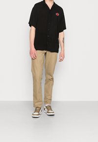 Camicetta nera con maniche corte e bottoni, con logo sulla tasca, abbinata a pantaloni beige e sneakers marroni e bianchi, in piedi su un pavimento grigio.