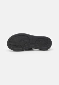 Semelle de chaussure en caoutchouc noir avec des rainures courbes et des motifs ondulés, vue de dessous sur un fond blanc.
