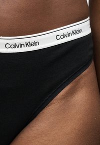 Κοντινό πλάνο της μέσης μαύρου εσώρουχου Calvin Klein σε γοφό και κάτω κορμό ατόμου με σκούρο δέρμα.