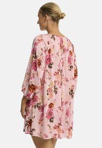 Modelo vista desde atrás, vistiendo un vestido floral rosa con mangas largas, que exhibe flores vibrantes contra un fondo claro.