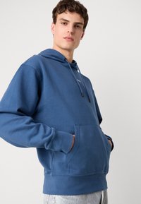 Hoodie in cotone blu con tasca frontale, cappuccio regolabile con cordino e polsini a coste. Presenta un design semplice con un branding minimo.