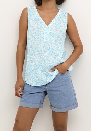 Personne portant un haut sans manches bleu et blanc à motif floral et un short en jean clair retroussé, avec une main dans la poche et un tatouage d’étoile sur la jambe gauche.