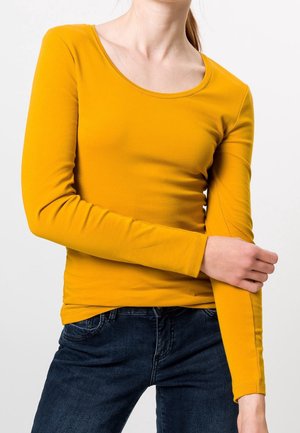 Osoba ubrana w musztardowo-żółtą bluzkę z długim rękawem i ciemnoniebieskie dżinsy, poprawiająca rękaw jedną ręką na neutralnym tle.