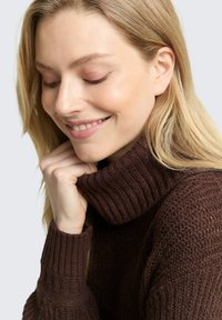 Brauner gestrickter Pullover mit hohem Rippenkragen und strukturiertem Muster. Das Haar ist hell und wellig, die Hand liegt auf dem Kragen.
