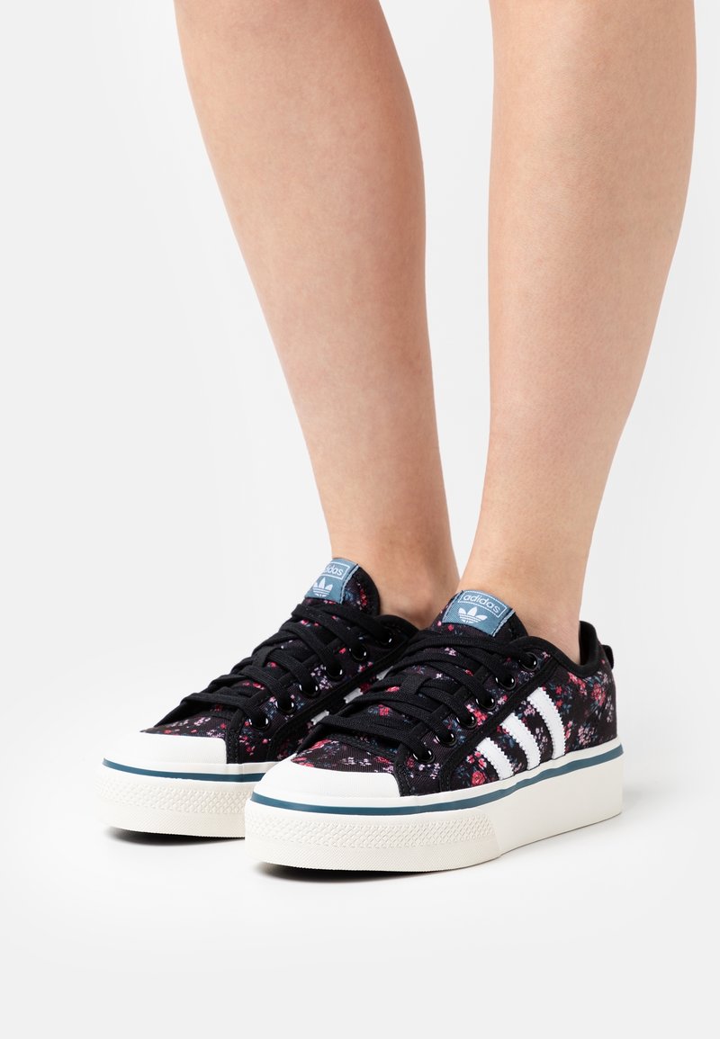 Svarta sneakers med blommönster, vita tre ränder, blå detaljer och en texturerad gummisula. Snörning och lågskuren design.