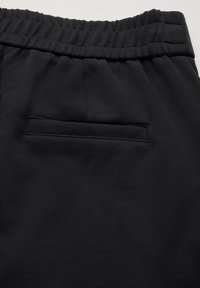 Pantalons noirs en tissu lisse, avec une taille élastique et une poche arrière cousue. La texture semble douce et décontractée.