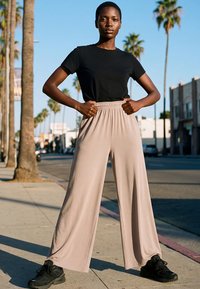 Kvinde står selvsikkert på en solrig gade med hænderne på hofterne, iført sort t-shirt, brede beige bukser og sorte sneakers.