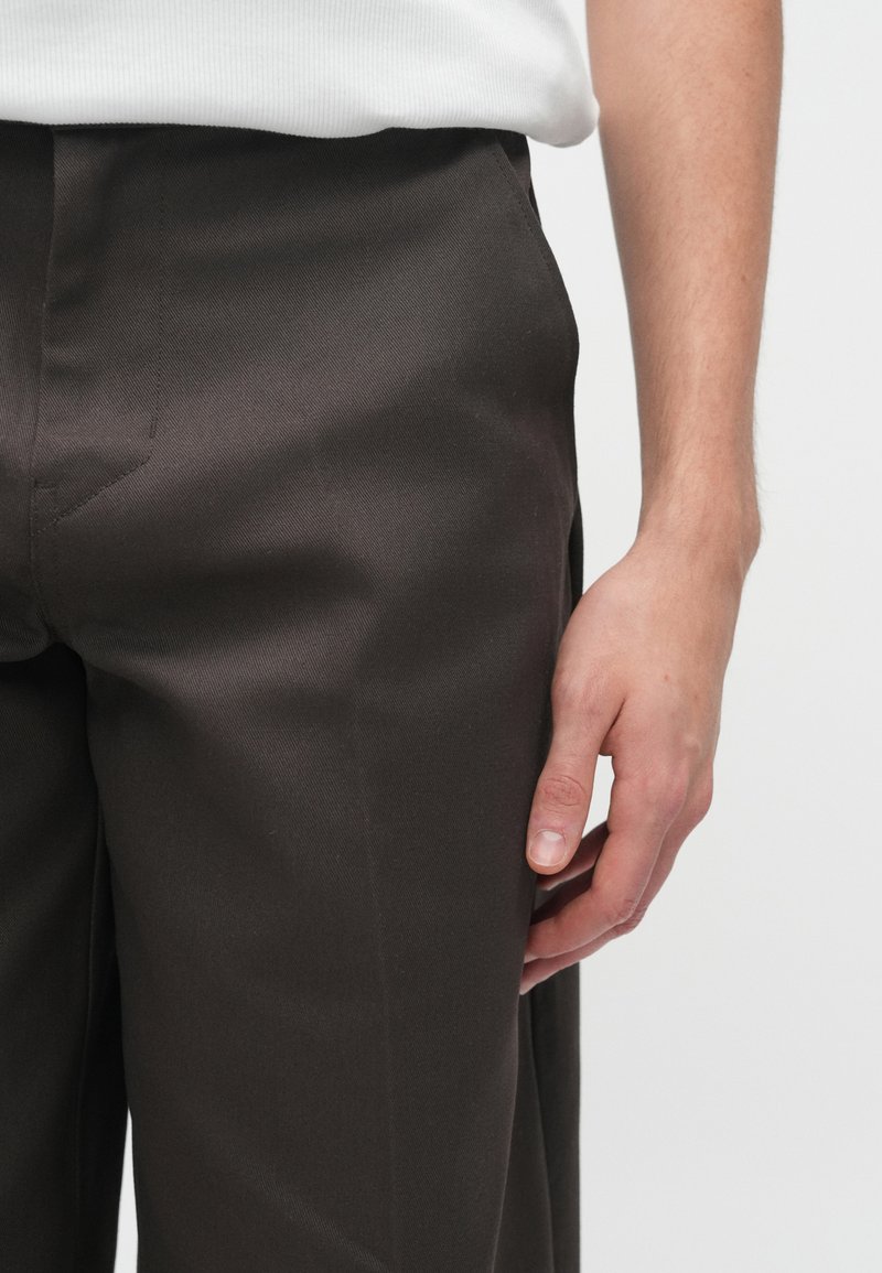 Brune skræddersyede bukser i et glat stof, med lige snit og subtil folder fortil, kombineret med en hvid top og en hånd på taljen.