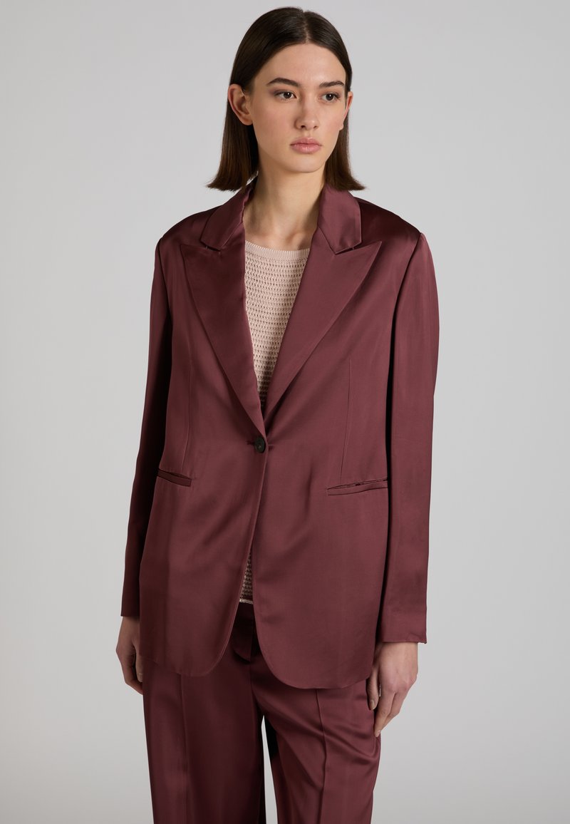 Junge Frau trägt ein burgunderrotes Satin-Blazer und passende Hose über einem beigefarbenen Stricktop und steht vor einem schlichten grauen Hintergrund.