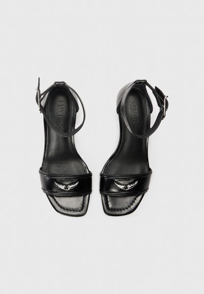 Čierne kožené sandále na vysokom opätku s remienkom okolo členka, otvorenou špičkou, širokými prednými pásmi a striebornými dekoratívnymi prvkami. Dizajn s plochou podrážkou.