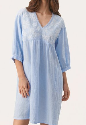 Femme portant une robe bleu clair jusqu'aux genoux avec des manches trois-quarts et une broderie florale blanche sur la poitrine.
