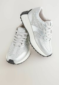 Srebrne sneakersy z gładką metaliczną cholewką, błyszczącymi kryształkami, wyściełanym językiem oraz czarną gumową podeszwą dla lepszej przyczepności.