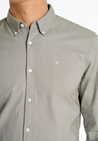 Camicia grigia chiaro con colletto, con bottoni bianchi e un piccolo logo ricamato sul petto. Texture liscia.