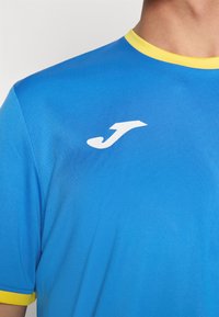 Blue athletic shirt made of smooth fabric, featuring a round yellow collar and a white logo on the chest. Short sleeves.