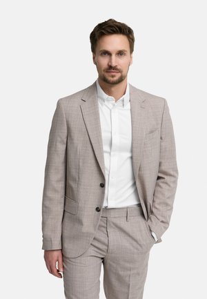 Hombre con cabello corto y castaño y barba, vestido con un traje beige claro y camisa blanca, de pie con una mano en el bolsillo.