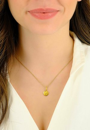 Collana con ciondolo in oro su una catena sottile, con un charm circolare decorato a rilievo. Indossata sopra una camicia bianca con colletto.