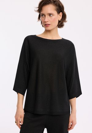 Kobieta o krótkich włosach, ubrana w luźną czarną bluzkę z rękawem 3/4 oraz dopasowane czarne spodnie, patrząca na bok na jednolitym tle.